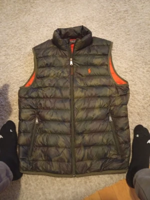 Camouflage dunväst från Polo Ralph Lauren - Säljer en dunväst från Polo Ralph Lauren i camouflage med orange insida och broderad logga på bröstet. Västen har hög krage, två fickor med dragkedja och quiltad design. Perfekt för lager-på-lager och riktigt snygg till hoodie eller tröja.