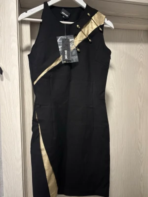 Svart och guld kortklänning Just Cavalli - Säljer en svart kortklänning från Just Cavalli med coola gulddetaljer och dekorativa nitar framtill. Klänningen är ärmlös och har en asymmetrisk guldpanel som ger en edgy look. Perfekt för dig som vill sticka ut med en unik design. Ordinarie pris, 5000kr