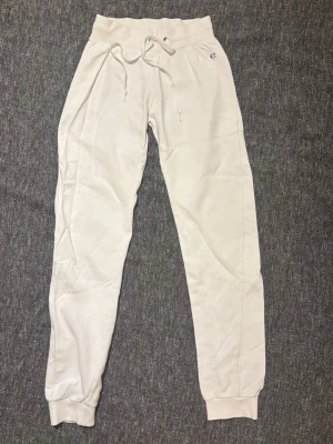 Vita mjukisbyxor från Champion S - Vita lågmidjade mjukisbyxor från Champion. Byxorna har en liten broderad Champion-logga vid höften och är gjorda i mjuk bomullsmix. Perfekta för chill dagar eller träning. Finns även en svag blå fläck vid slutet av byxan.