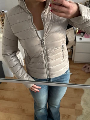 Beige pufferjacka från Mango - Snygg beige pufferjacka med horisontella quiltade sömmar och två fickor med dragkedja. Jackan har hög krage och hel dragkedja framtill. Materialet är glansigt och känns lätt, perfekt för kyliga dagar. Passformen är lite liten i storlek, sitter som S. 💕💕Endast använd ett fåtal gånger