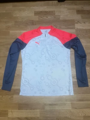 Puma future tracksuit - Bilderna visar ett träningsset från Puma med långärmad tröja och byxor, men inga skor finns med. Tröjan är vit med mönster, korallröda axlar och mörkblå ärmar. Byxorna är mörkblå med korallröda och vita detaljer. Storlek L.