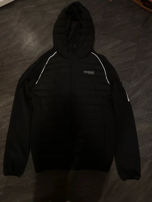 Svart pufferjacka från McKenzie - Svart pufferjacka från McKenzie med huva och vita detaljer längs axlarna. Jackan har quiltad framsida, dragkedja och två sidofickor. Perfekt för dig som vill ha en stilren och sportig look under kyliga dagar.