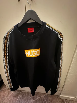Svart sweatshirt från Hugo Boss M - Svart långärmad sweatshirt från Hugo Boss med gul och vit logga på bröstet. Snygga band med HUGO-tryck i vitt och gult längs ärmarna. Rund hals och normal passform. Materialet är mjuk bomull, perfekt för chill dagar.