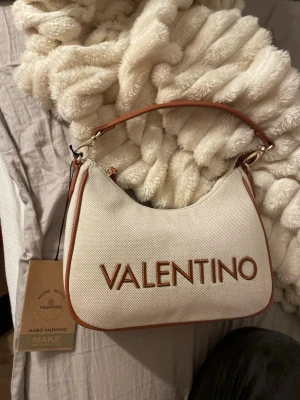 Beige axelväska från Valentino - Snygg beige axelväska från Valentino med bruna detaljer och stort broderat logotyp framtill. Väskan är tillverkad i kraftig bomull med bruna skinnpartier och har en praktisk dragkedja upptill. Insidan har flera fack för dina grejer. Perfekt för dig som gillar stilren design.