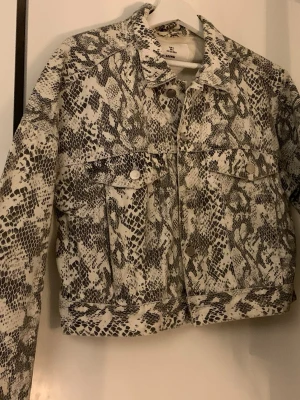 Jeansjacka med ormmönster från H&M - Säljer en jacka från H&M med ormmönster i svart och vitt (grått). Jackan har klassisk jeansjacke-modell med knappar framtill och två bröstfickor. 