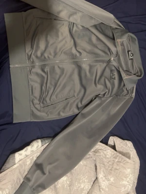 Grå bomberjacka från EA7 Emporio Armani - Snygg grå bomberjacka från EA7 Emporio Armani med dragkedja framtill och två fickor. Jackan har ribbade muddar vid ärmslut och midja samt diskret logga på bröstet. Perfekt för dig som gillar stilren och sportig look.