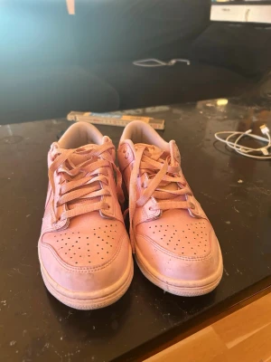 Nike Dunk Low rosa sneakers - Säljer ett par Nike Dunk Low sneakers i en snygg ljusrosa färg. Skorna har klassisk snörning, perforerad tå och den ikoniska swoosh-loggan på sidan. Tillverkade i skinn och syntetmaterial för en clean look. Perfekta för dig som vill sticka ut med stil.