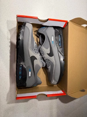 Nike Air Max 90 grå blåa sneakers - Säljer ett par Nike Air Max 90 sneakers i grått med svarta detaljer och blå logga. Skorna har en klassisk siluett med mesh och syntetmaterial, synlig Air-enhet i sulan och snörning. Perfekta för dig som gillar stilrena och sportiga sneakers. Kostar helt nya 1900kr mina är helt nya aldrig använda