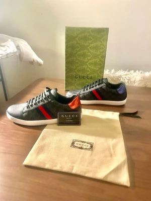 Gucci sneakers med röd och blå detalj - Säljer ett par svarta Gucci sneakers med klassiskt GG-mönster, röda och blå ränder på sidan samt metallicröd detalj på ena hälen och metallicblå på den andra. Skorna har snörning, vit platt sula och är tillverkade i skinn och syntet. Box och dustbag medföljer.