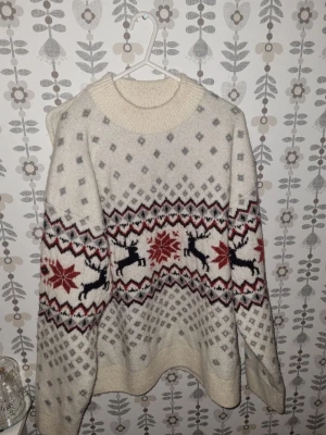 Vit stickad jultröja med renar - Mysig vit stickad tröja med grått och rött mönster, renar och snöflingor. Tröjan har rund hals och långa ärmar, perfekt för vintern och julen. Den är oversized och har en klassisk nordisk look.