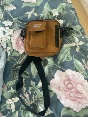 Brun Carhartt axelväska - Säljer en brun axelväska från Carhartt med svart justerbar rem. Väskan har en praktisk ficka framtill med lock och en sidoficka med dragkedja. Perfekt för att bära med sig det viktigaste och har en stilren design med Carhartt-logga på framsidan.