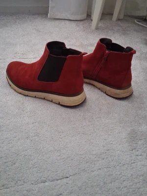 Röda chelsea boots från Tamaris - Säljer ett par snygga röda chelsea boots från Tamaris i storlek 38. Skorna är i mocka med elastiska svarta paneler på sidorna och dragkedja på insidan. De har en beige, räfflad sula och rund tå. Perfekta för dig som vill ha ett statement-plagg till hösten!