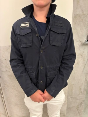 Field jacket  - Snygg mörkblå jacka med militärvibbar, flera fickor framtill och coola patchar på bröstet och ärmen. Jackan har hög krage, knappar hela vägen och är tillverkad i slitstark bomull. Stilren och snygg, hör av er vid frågor!