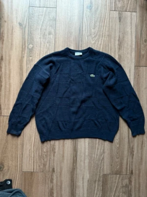 Mörkblå stickad tröja från Lacoste - Säljer en mörkblå stickad tröja från Lacoste med klassisk rund hals och broderad krokodil-logga på bröstet. Tröjan har ett snyggt rutmönster i stickningen och är tillverkad i en mix av ull och akryl. Är i storlek M men passar mer som L.  Pris kan såklart diskuteras.