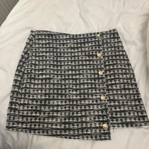 Rutig kortkjol från Vero Moda - Säljer en svartvit rutig kortkjol från Vero Moda med snygga guldknappar längs framsidan. Kjolen har en klassisk A-linjeform och är tillverkad i ett strukturerat tyg som ger en trendig look.