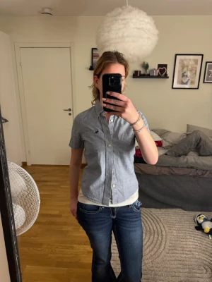 Abercrombie & Fitch Skjorta - Snygg kortärmad skjorta från Abercrombie & Fitch i en fräsch blågrå "chambray"-färg. Det är en barnstorlek XL, vilket motsvarar ungefär en vuxen XS (se bild för passform). Modellen är "Muscle Fit", vilket ger en snygg och figurnära siluett. Perfekt för sommarens alla tillfällen! • Märke: Abercrombie & Fitch • Storlek: Kids XL (Passar vuxen XS) • Modell: Muscle Fit • Skick: Toppskick • Nypris: Ca 450–600 kr.