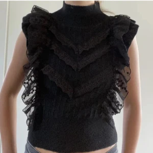 Svart volang topp med spetsdetaljer - Supersnygg svart topp med höghalsad design och volanger i lager på framsidan. Spetsdetaljer längs volangerna ger en cool och unik look. Toppen är ärmlös och croppad, perfekt för att sticka ut med en edgy vibe.