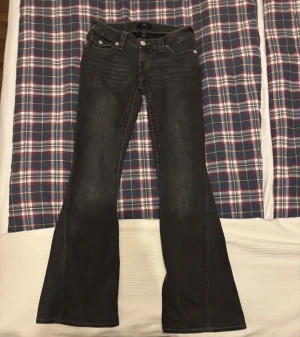 True Religion Joey Low Rise Flare Jeans - Snygga svarta jeans från True Religion, modell Joey Low Rise Flare. De har låg midja, utställda ben och coola detaljer med silverfärgade knappar på bakfickorna. Jeansen är tillverkade i stretchigt denim och har klassisk femficksdesign.