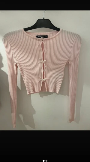 Ljusrosa knyten ribbad topp - Superfin ljusrosa croppad toppmed ribbad struktur och långa ärmar. Toppen har tre söta rosetter framtill och är tillverkad i mjuk bomullsmix. Perfekt för dig som gillar gulliga detaljer och en feminin look.