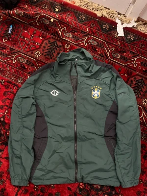 1 OF 1 Grön vindjacka med Brasilienmärke - Grön vindjacka med svarta paneler och dragkedja framtill. Jackan har Brasilien-emblem på bröstet och tryck på ryggen. Hög krage och elastiska muddar. Perfekt för dig som gillar sportig stil och vill sticka ut med snygga detaljer.