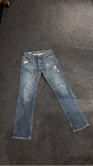 Blå jeans från Abercrombie & Fitch - Snygga blå jeans från Abercrombie & Fitch med klassisk femficksdesign och slitna detaljer på fram- och bakfickor. Jeansen har raka ben, knappgylf och kontrastsömmar. Perfekt för dig som gillar en avslappnad och cool stil.
