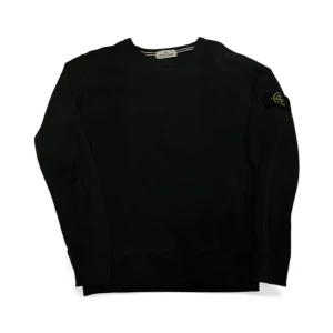 Svart Stone Island sweatshirt - Svart sweatshirt från Stone Island med klassisk rund hals och ribbade muddar. Tröjan har lång ärm och ikonisk logotyp-patch på vänster ärm. Tillverkad i mjuk bomull för skön komfort och clean look. Perfekt för dig som gillar streetwear och stilrena plagg.