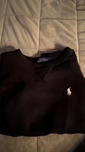 Svart sweatshirt från Polo Ralph Lauren XS - Svart sweatshirt från Polo Ralph Lauren i storlek XS. Klassisk rund hals och långärmad modell med den ikoniska vita Polo-loggan broderad på bröstet. Mjuk bomullskvalitet och ribbade muddar vid ärmslut och nederkant. Perfekt för en chill och stilren look.