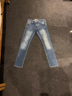 Blå jeans från Visual Clothing Project - Snygga blå jeans från Visual Clothing Project i storlek 32. Modellen har klassisk femficksdesign, ljusa slitningar framtill och raka ben. Jeansen är tillverkade i mjukt denim med kontrastsömmar och knappstängning i midjan.