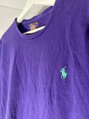 Lila t-shirt från Polo Ralph Lauren - Snygg lila t-shirt från Polo Ralph Lauren i slim fit-modell. Klassisk rund hals och korta ärmar. T-shirten är i mjuk bomull och har den ikoniska gröna Polo-loggan broderad på bröstet. Perfekt för dig som gillar stilrena och färgstarka plagg.