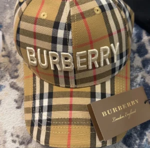 Burberry rutig keps med broderi - Snygg keps från Burberry med klassiskt rutig mönster i beige, svart, vitt och rött. Stor broderad Burberry-logga framtill. Kepsen har böjd skärm och justerbar passform. Perfekt för dig som vill ha en ikonisk streetstil med lyxig touch.