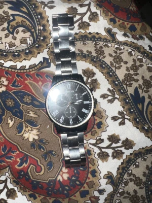 Svart och silver klocka från Geneva - Stilren klocka från Geneva med svart urtavla och romerska siffror i vitt. Armbandet är i blankt silverfärgat metall och har en klassisk länkdesign. Urtavlan har tre dekorativa subdials och tydliga detaljer. Perfekt accessoar för dig som gillar en clean look.