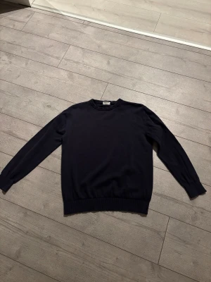 Mörkblå stickad tröja från Kappahl  - En stilren mörkblå stickad tröja från Kappahl med rund hals och ribbade muddar vid ärmslut och nederkant. Perfekt för dig som gillar klassisk och enkel design. Tröjan har lång ärm och är gjord i mjukt material som känns skönt mot huden.