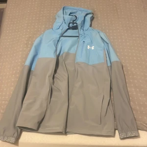 Blå och grå vindjacka från Under Armour - Snygg tvåfärgad vindjacka från Under Armour i ljusblått och grått. Jackan har huva, dragkedja framtill, lång ärm och logga på bröstet. Perfekt för blåsiga dagar och sportiga outfits. Materialet är lätt och känns smidigt mot kroppen.