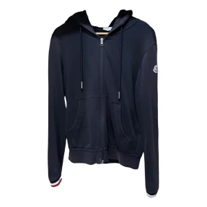 Mörkblå hoodie från Moncler - Snygg mörkblå hoodie från Moncler med dragkedja, huva och snörning. Tröjan har två fickor fram och Moncler-logga på ärmen. Muddarna har röd, vit och blå detalj. Perfekt för dig som gillar stilrena och exklusiva plagg.