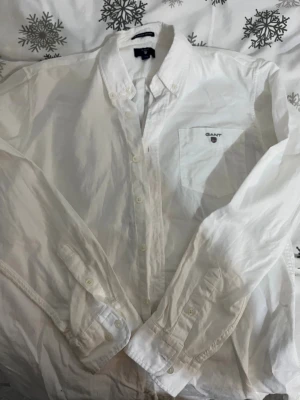 Vit skjorta från GANT med knapp - Vit klassisk skjorta från GANT med knappar hela vägen och bröstficka med broderad logga. Skjortan har lång ärm, button-down krage och är gjord i mjuk bomull. Perfekt för en clean och stilren look.
