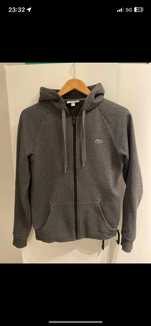 Grå hoodie från Lacoste med dragkedja - Snygg mörkgrå hoodie från Lacoste med hel dragkedja och huva med snörning. Klassisk krokodillogo på bröstet och tryck på huvan. Mjuk bomullsmix, två fickor fram och långärmad modell. Perfekt för chill dagar eller när du vill ha en clean look.
