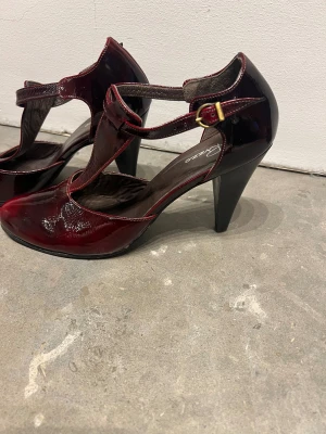 Vinröda lackpumps från Rizzo - Snygga vinröda pumps från Rizzo i glansigt lackskinn med rund tå och hög klack. Skorna har ett elegant T-rem med guldfärgat spänne och fina söm-detaljer. Perfekt för dig som gillar att sticka ut med stilrena och trendiga skor.