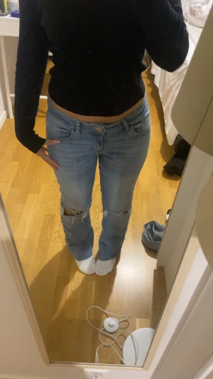 Ljusblå ripped jeans med låg midja - Säljer ett par ljusblå jeans med låg midja och utsvängda ben. De har slitna hål vid knäna för en cool, avslappnad vibe. Klassisk femficksmodell och knappstängning framtill. Perfekta för dig som gillar streetstyle och vill ha en chill look.