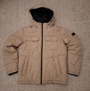 Beige pufferjacka med avtagbar huva - !Läs Hela Beskrivningen! Säljer en beige pufferjacka från jack & Jones i storlek L. Jackan har avtagbar huva, två stora bröstfickor med knapp, sidofickor och svart innerfoder. Stängs med dragkedja framtill och har en clean, boxig look som är perfekt för kalla dagar, perfekt inför den kalla vintern. !Det finns en fläck på högra axeln av jackan som visas på sista bilden i annonsen! Priset kan disluteras.