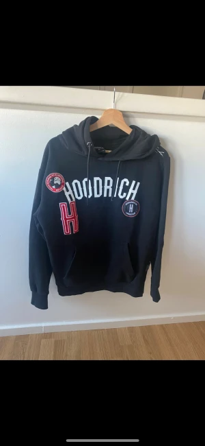 Svart hoodie från Hoodrich - Svart hoodie från Hoodrich med stora broderade patchar och tryck i vitt och rött framtill. Klassisk huva med snörning och känguruficka. Perfekt för dig som gillar streetwear och vill sticka ut med detaljerade loggor.