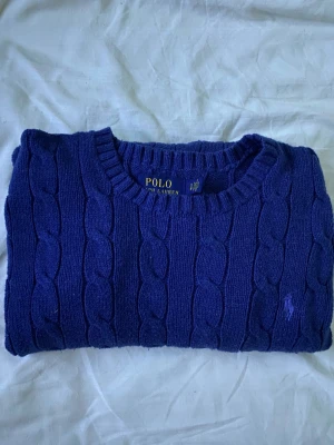 Blå stickad tröja från Polo Ralph Lauren - Snygg blå stickad tröja från Polo Ralph Lauren med klassiskt kabelstickat mönster och rund hals. Tröjan har lång ärm och den ikoniska broderade loggan på bröstet. Perfekt för dig som gillar preppy stil och vill ha något som känns både chill och stilrent. Fint skick, lite liten i storleken men den är stretchig. Kan gå ner i pris vid snabb affär! 