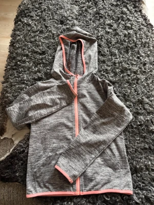 Grå träningsjacka med aprikosa detaljer - Sportig grå vindjacka med huva och hel dragkedja i rosa. Jackan har ett diskret mönster, praktiska fickor och rosa kantband längs ärmar och nederkant. Perfekt för träning eller chill dagar. Snygg och funktionell design med lång ärm och mjukt syntetmaterial.