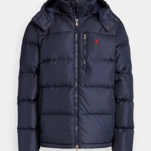 Mörkblå pufferjacka från Polo Ralph Lauren - Snygg mörkblå pufferjacka från Polo Ralph Lauren med huva och dragkedja framtill. Jackan har en röd logga på bröstet, praktiska fickor och quiltad design som ger en cool look. Perfekt för kalla dagar och enkel att matcha med din stil.