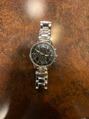 Stilren silverfärgad kronografklocka - Snygg kronografklocka med svart urtavla och flera små visare. Armbandet är i blankt silverfärgat metall med robust länkkonstruktion och smidig låsning. Klockan har rund form och detaljerade markeringar på urtavlan, perfekt för dig som gillar klassisk men modern stil. 14-17Cm