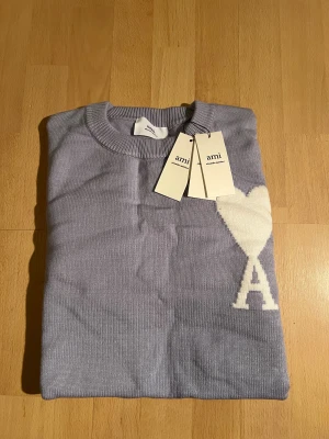 Ljusblå tröja från Amiparis - Snygg ljusblå stickad tröja från Ami Alexandre Mattiussi med rund hals och ribbade muddar. På bröstet finns ett vitt hjärta med bokstaven A. Perfekt för dig som gillar stilrena detaljer och mjuka material.