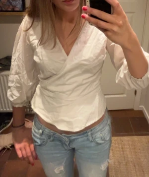 Vit omlottblus med trekvartsärm - Säljer en fräsch vit omlottblus med trekvartsärmar och lätt puff vid axlarna. Blusen har v-ringning och en figurnära passform som framhäver midjan. Perfekt att styla med jeans eller kjol för en clean look.