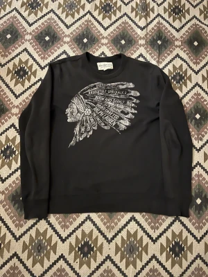 Svart sweatshirt från Ralph Lauren - Svart sweatshirt från Denim & Supply Ralph Lauren med stort tryck framtill av ett indianskt huvudbonadsmotiv och text. Tröjan har rund hals, långa ärmar och är tillverkad i mjuk bomull. Perfekt för dig som gillar streetwear och coola prints.