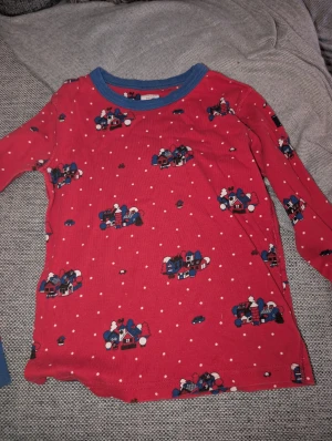 Röd långärmad tröja Polarn O. Pyret stl 122-128 - Säljer en röd långärmad tröja från Polarn O. Pyret med blå kant vid halsen. Tröjan har ett gulligt vintermönster med små hus och snöprickar över hela plagget. Tillverkad i mjuk bomull, perfekt för kalla dagar och mysiga stunder.