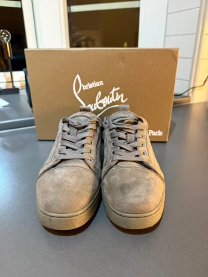 Grå mocka sneakers Christian Louboutin - Säljer ett par gråa sneakers från Christian Louboutin i mjuk mocka med klassisk rund tå och platt sula. Skorna har snörning och den ikoniska röda sulan med Louboutin-loggan. Perfekt för dig som vill ha en clean och lyxig look.