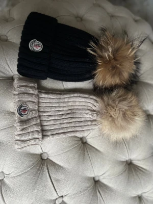 Moncler mössa med pälsboll - Två ribbstickade mössor från Moncler, en svart och en beige, båda med stor fluffig pälsboll i bruna toner på toppen. Mössorna har Moncler-logga framtill och är tillverkade i mjukt stickat material. Perfekt för dig som vill ha en snygg och varm mössa i vinter. Pälsen är även avtagbar säljer för 200kr st men pris kan såklart diskuteras båda är i nyskick och Aldrig använda .